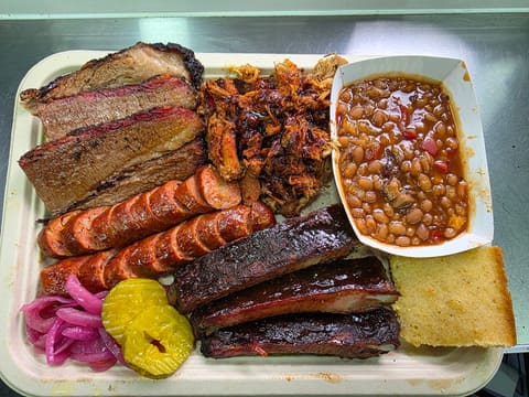 An overhead view of a loaded BBQ combo tray: sliced brisket, chopped pulled pork, smoked sausage, glazed ribs, baked beans, cornbread and pickles.