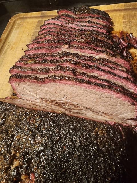 Close-up of smoked beef brisket sliced on a wooden board, showing a thick black pepper bark and a deep pink smoke ring.