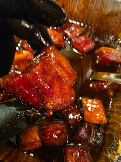 A gloved hand lifting a lacquered burnt end from a pan of sauce, the cube glazed deep red and dripping.