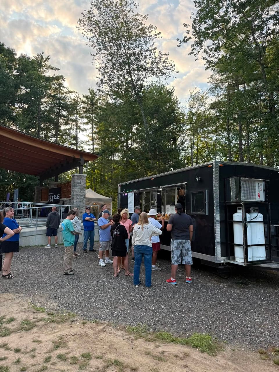 About a dozen customers queued at the Gator Pit BBQ trailer's serving window on a gravel lot at golden hour, with an outdoor pavilion and tall pines behind them.