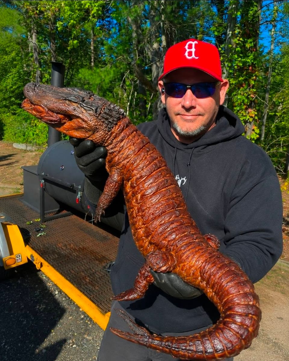 The pitmaster beside his offset smoker in the New Hampshire woods, holding up a whole smoked alligator with a deep mahogany lacquered hide.