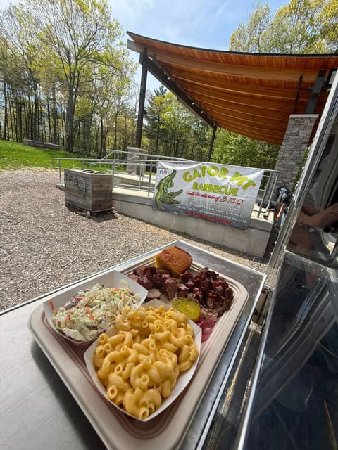 A BBQ plate of macaroni and cheese, coleslaw, sliced sausage, chopped pork, cornbread and a pickle on the trailer's stainless serving counter at an outdoor event.
