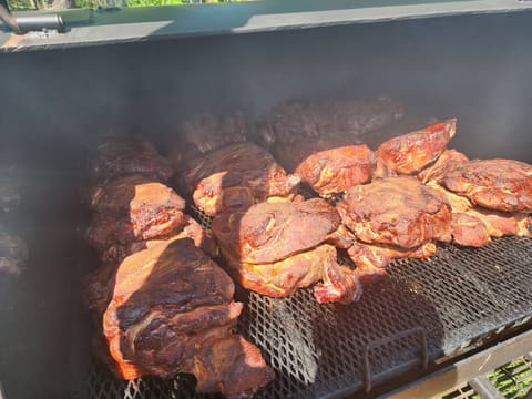 A dozen pork shoulders on the smoker grate in mid-cook, mahogany and rust coloured, with sunlit smoke drifting through the chamber.