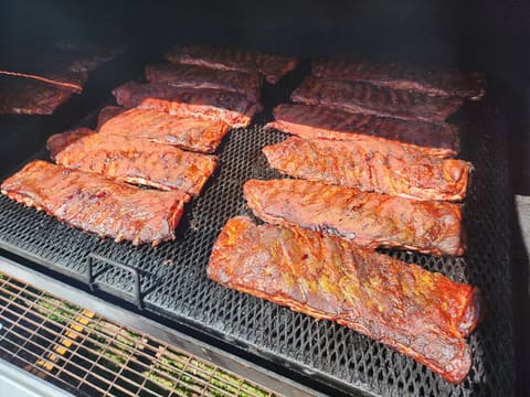 A dozen full racks of pork ribs laid across the diamond-plate grate inside the smoker, glistening mahogany red under a haze of smoke.
