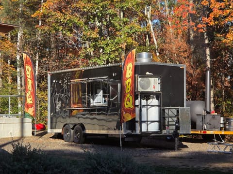 The black Gator Pit BBQ trailer parked on gravel in front of blazing orange New Hampshire fall foliage, with the offset smoker hitched behind it.