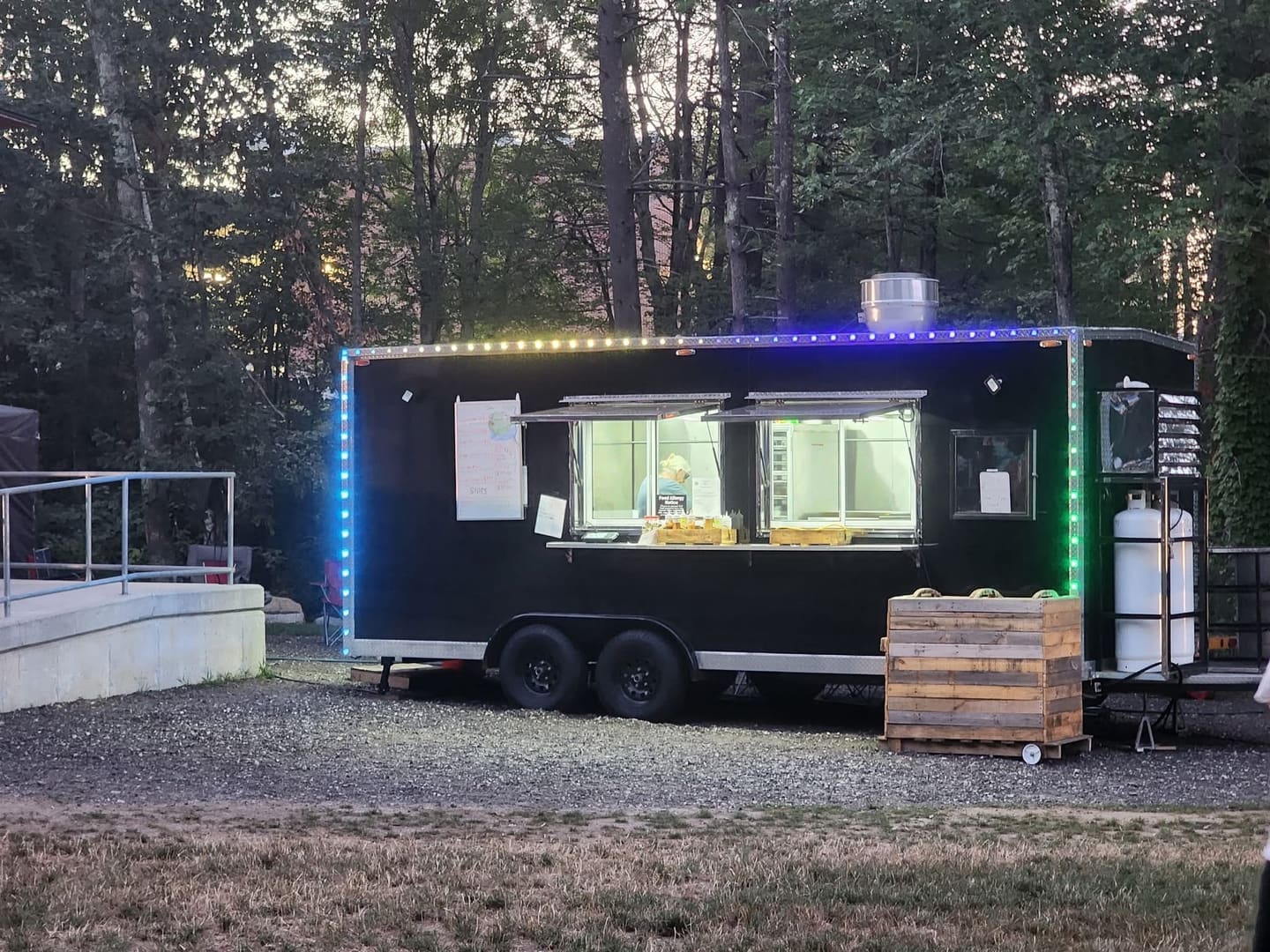 The Gator Pit BBQ trailer at dusk with its serving window open and LED rope lights outlining the roof, a staff member working inside the lit galley.